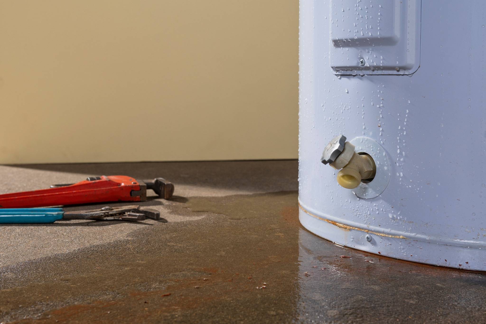 A burst pipe near a water heater causes leaking water to pool on the floor beside plumbing tools.