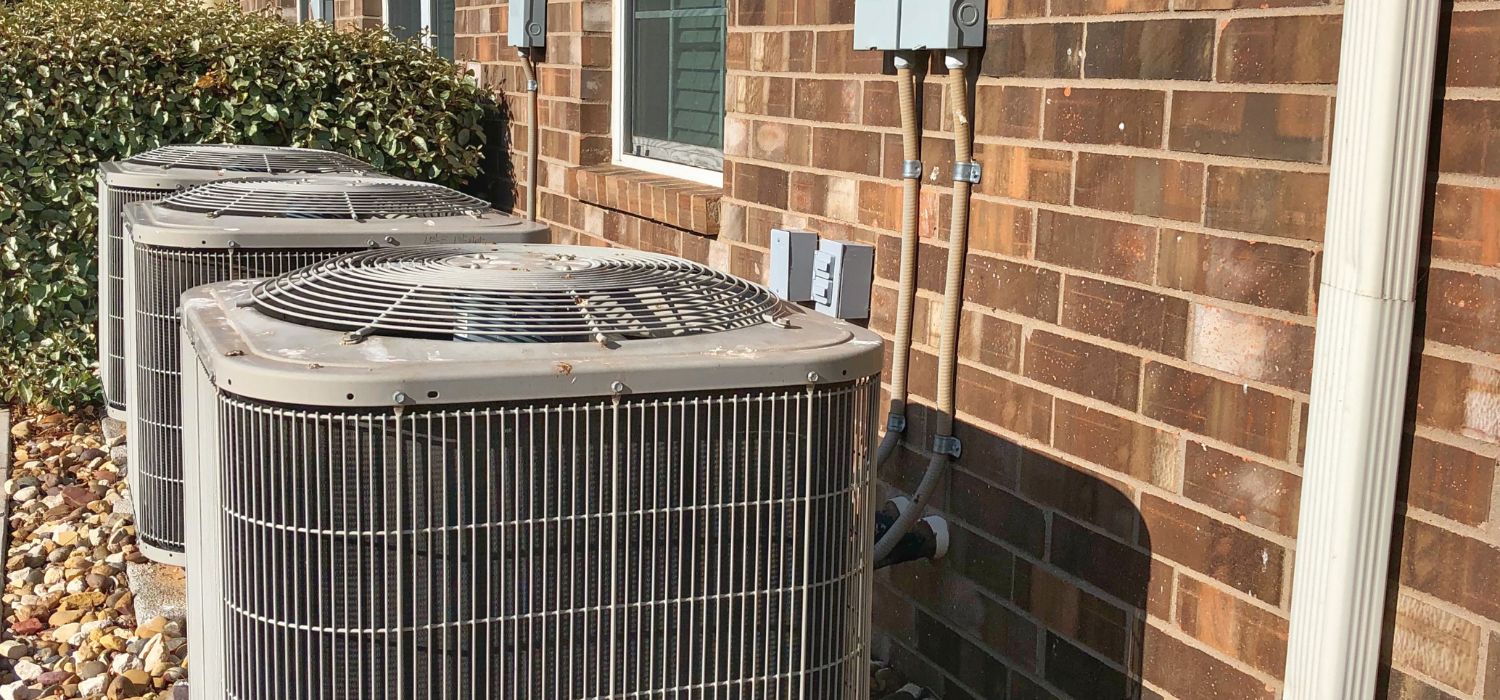 Two outdoor HVAC cooling units installed beside a brick building, with electrical connections and landscaping visible in a desert climate setting.